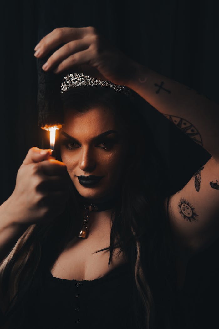 Dark portrait of a tattooed woman lighting a candle, showcasing gothic style and black lipstick.