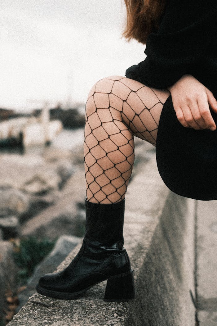 A fashionable woman in fishnet stockings sits on a seaside wall wearing stylish black boots.